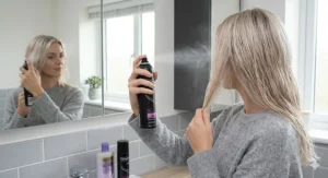 A person applying heat protection spray to damp blonde hair before using a hairdryer to prevent breakage.