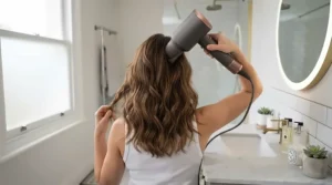 A woman viewed from behind, holding the hairdryer at a high angle to style the crown of her head, with the swivel cord moving freely over her shoulder.